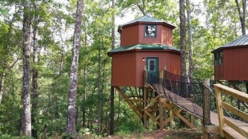 A red treehouse perched among trees, connected by a wooden bridge, surrounded by lush greenery.