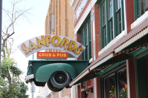 Sign for Sanford's Grub & Pub featuring a truck bed design, hanging above a brick building in a city setting.