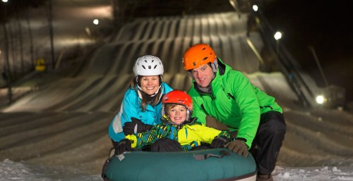 A family smiles while sitting on a snow tube at a winter tubing hill, wearing helmets and colorful jackets.