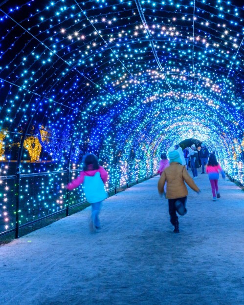 Children run through a brightly lit tunnel of blue and white lights, with adults walking in the background.