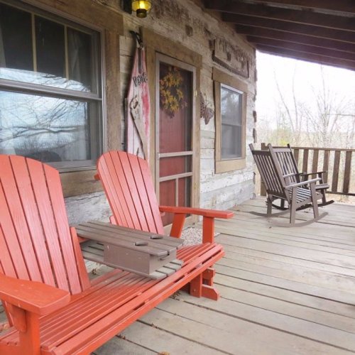 Two red Adirondack chairs and a rocking chair on a rustic wooden porch with a view of trees outside.