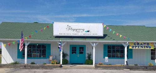 A storefront with a green roof, blue accents, and a sign reading "Flipping" with colorful flags and a "Grand Opening" banner.
