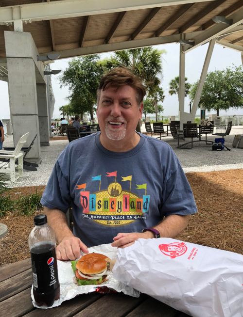 A man with a Disney-themed shirt sits at a table outdoors, enjoying a burger and a drink.
