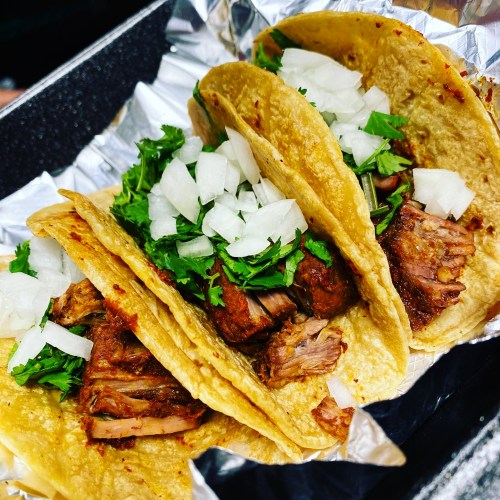 Three tacos filled with tender meat, topped with chopped onions and fresh cilantro, served in a foil-lined container.