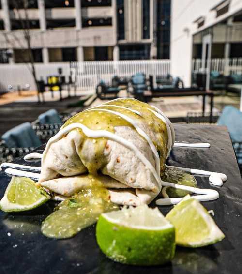 A burrito drizzled with green sauce and sour cream, served with lime wedges on a black plate outdoors.