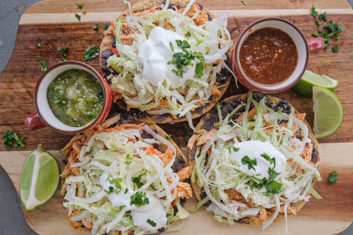 Three colorful tostadas topped with shredded lettuce, cheese, and sour cream, served with salsa and lime wedges.