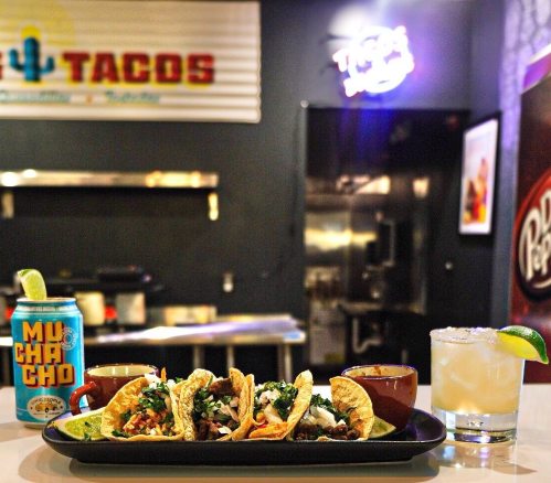 A platter of tacos with toppings, a drink can, and a cocktail on a table in a vibrant taco restaurant setting.