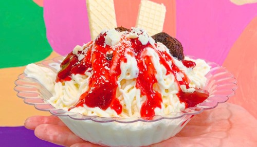 A hand holding a bowl of dessert topped with whipped cream, chocolate, and strawberry sauce, with wafer sticks.