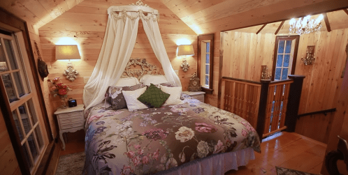 Cozy wooden bedroom with a floral bedspread, draped canopy, and warm lighting, featuring a staircase and decorative accents.