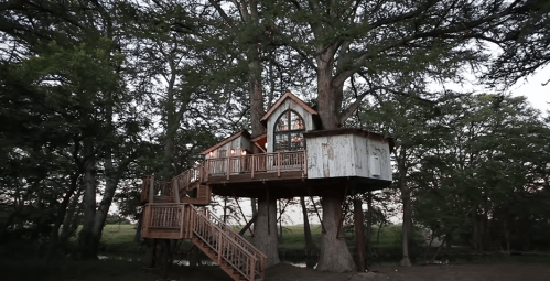 A charming treehouse with a wooden deck, nestled among trees, featuring large windows and a rustic design.