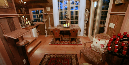 Cozy interior with wooden decor, a dining table, chairs, candles, and a patterned rug, illuminated by natural light.