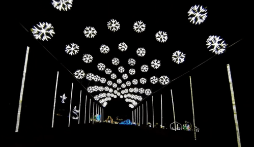A dark pathway illuminated by white snowflake lights hanging from above, creating a festive winter atmosphere.