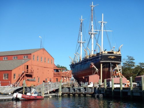 A historic ship docked at a harbor next to a red building under a clear blue sky.