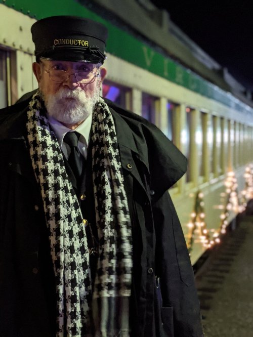 A conductor in a black coat and hat stands beside a decorated train at night, with festive lights in the background.