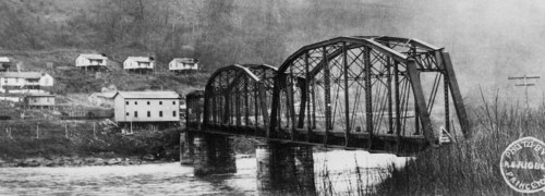 Black and white photo of a metal bridge over a river, with houses on a hillside in the background.