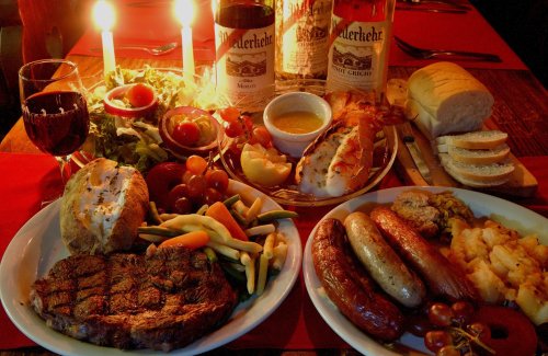 A lavish dinner spread featuring steak, sausages, seafood, vegetables, bread, and wine, set with candles.