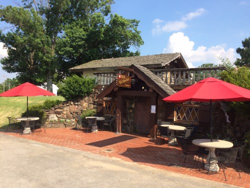 A rustic building with a stone facade, featuring outdoor seating under red umbrellas and a sign above the entrance.