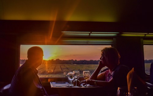 A couple sits at a table in a train, gazing out at a sunset over fields, with glasses of wine in front of them.