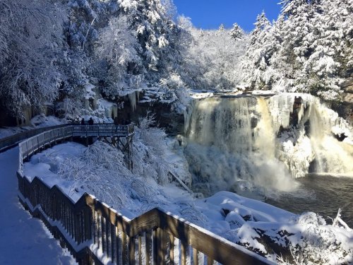 A snowy landscape featuring a waterfall, surrounded by trees and a wooden walkway under a clear blue sky.