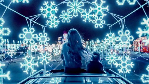 Two children sit on a car, surrounded by glowing blue snowflake lights in a festive winter display.
