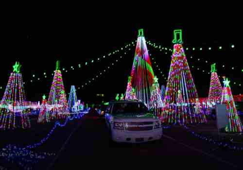 A festive display of colorful Christmas trees and lights, with a white vehicle in the foreground at night.