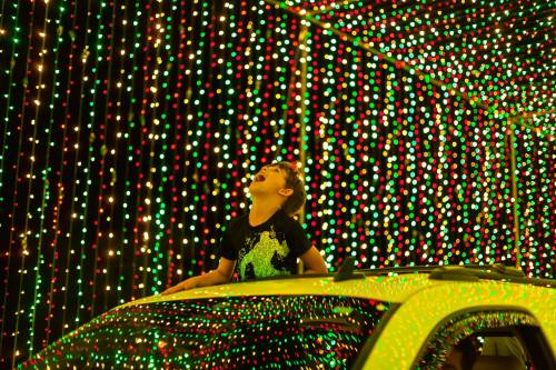 A child joyfully leans out of a car sunroof, surrounded by colorful holiday lights.