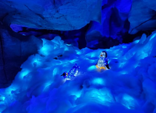 Two illuminated penguin figures on a bed of blue, glowing snow under a dark, icy cave.