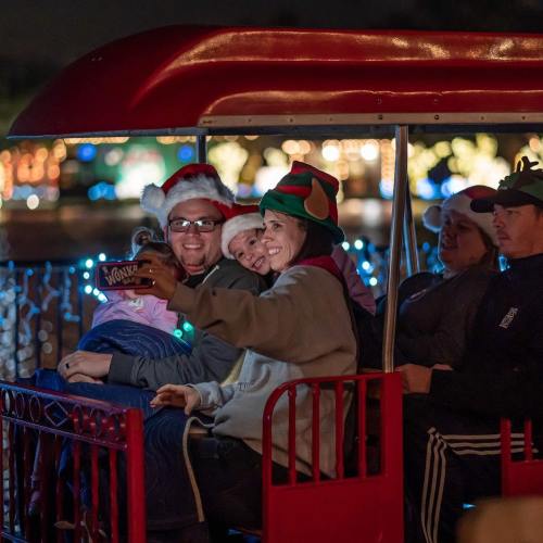 A family in festive hats takes a selfie on a decorated train ride at night, surrounded by colorful lights.