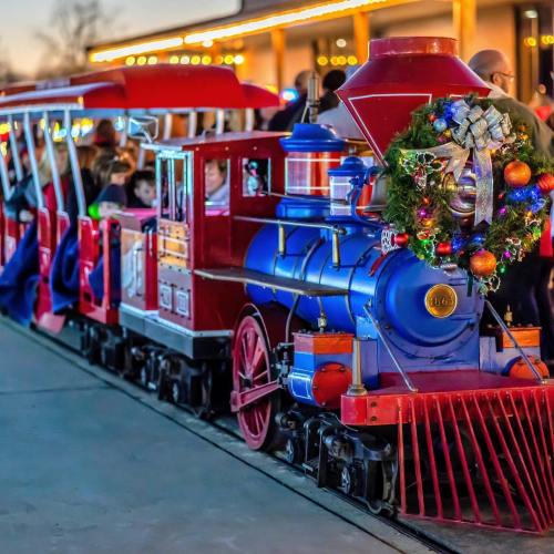 A colorful holiday train decorated with a wreath, surrounded by people in a festive setting.