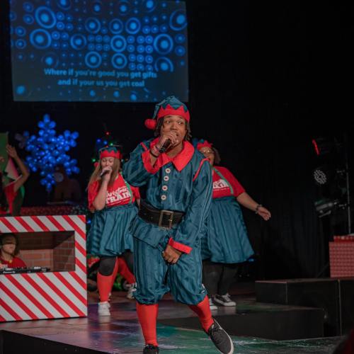 A performer in an elf costume sings on stage, surrounded by dancers in festive attire, with holiday decorations in the background.