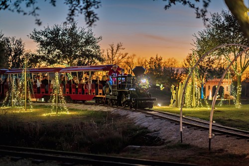 A festive train with passengers travels along a track, surrounded by twinkling lights at sunset.