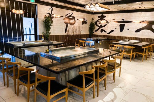 Interior of a modern restaurant featuring a large table with grills, wooden chairs, and decorative wall art.