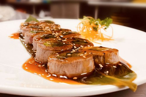 Sliced beef tenderloin drizzled with sauce, garnished with sesame seeds and a sprig of parsley on a white plate.
