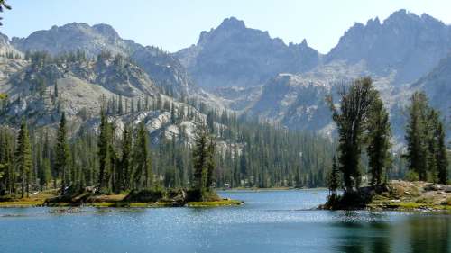Scenic view of a tranquil lake surrounded by tall trees and rugged mountains under a clear blue sky.
