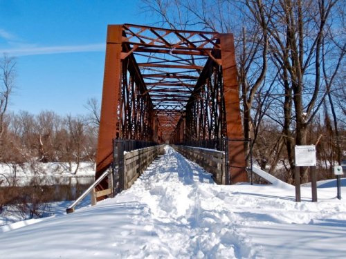 A snow-covered bridge with a metal frame spans a river, surrounded by bare trees under a clear blue sky.