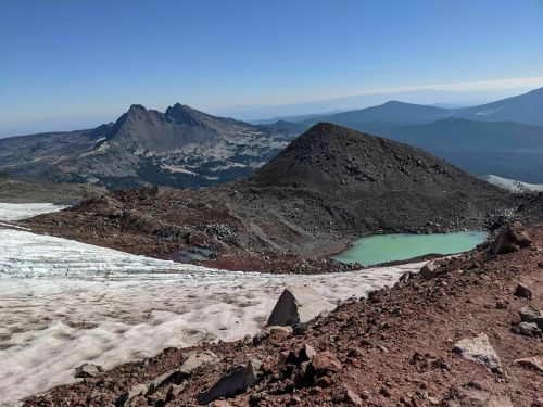 A panoramic view of rugged mountains and a greenish pond, with snow patches in the foreground under a clear blue sky.