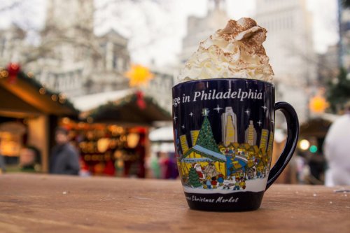 A festive mug with whipped cream and cocoa, set against a blurred holiday market in Philadelphia.