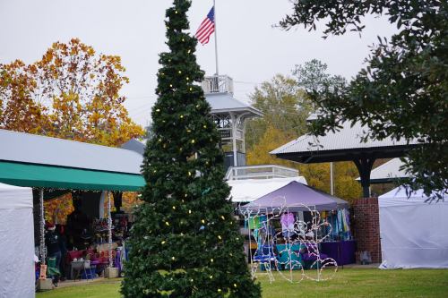 A decorated Christmas tree stands in front of vendor tents at a festive outdoor market, with an American flag in the background.