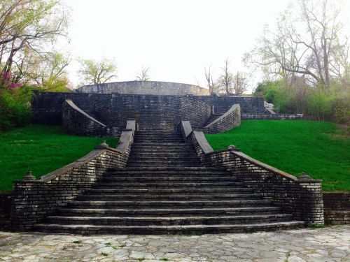 Stone steps lead up to a circular structure, surrounded by greenery and trees in a serene outdoor setting.