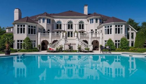 A luxurious mansion with large windows and a pool in the foreground, surrounded by lush greenery under a clear blue sky.
