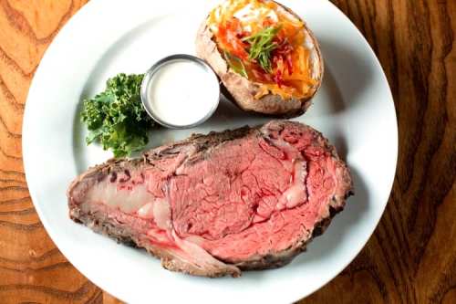 A plate featuring a juicy slice of prime rib, a baked potato with toppings, and a side of kale.