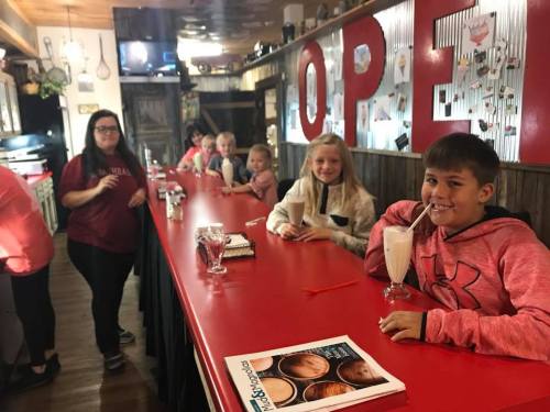 A group of children and a woman at a diner counter, enjoying milkshakes and chatting in a cozy setting.