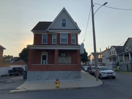 A two-story brick house on a street corner, with a fire hydrant in front and cars parked along the road.
