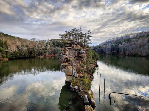 A rocky island with trees surrounded by calm water, reflecting a cloudy sky and distant hills.