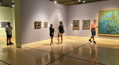 Visitors explore an art gallery, admiring paintings on the walls and reading information displays.