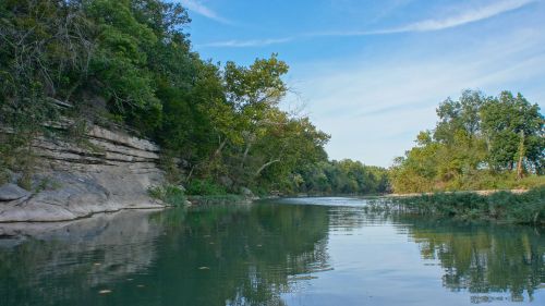A serene river flows between lush green trees and rocky banks under a clear blue sky.