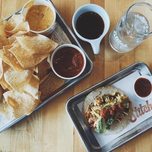 A tray with tortilla chips, cheese dip, salsa, and a taco topped with vegetables and sauce, alongside a glass of water.