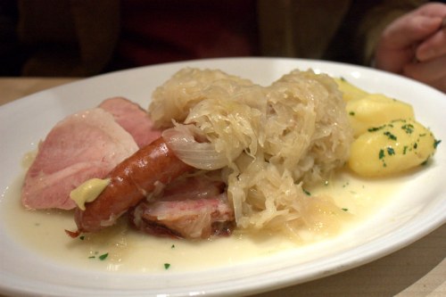 A plate of sauerkraut with slices of ham, sausage, and boiled potatoes, garnished with herbs.