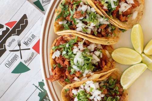 Three tacos filled with meat, topped with onions and cilantro, served with lime wedges on a plate.