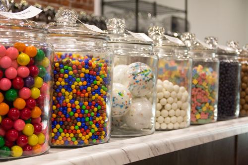 A row of glass jars filled with colorful candies, including gumballs, chocolate balls, and sprinkles.
